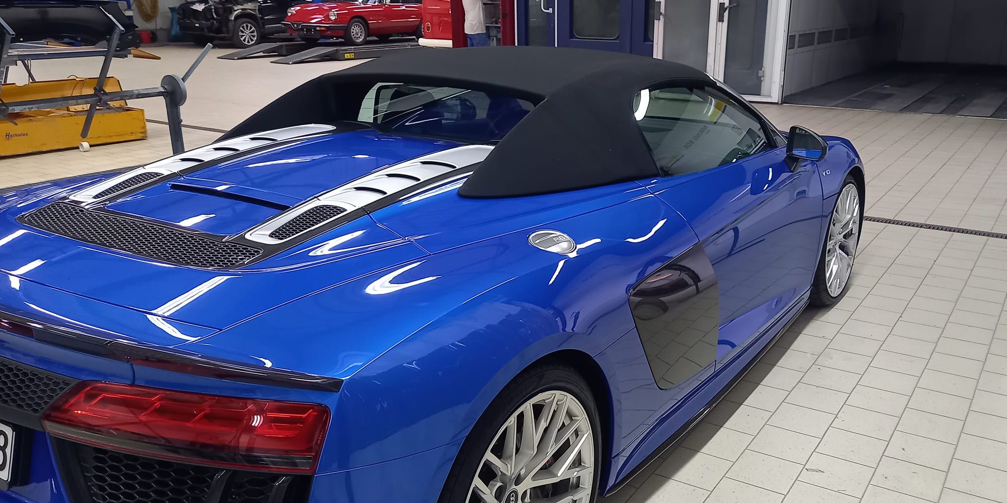 In einer Garage steht ein schnittiger blauer Sportwagen mit schwarzem Verdeck. Er hat eine glänzende Karosserie und markante Lüftungsschlitze